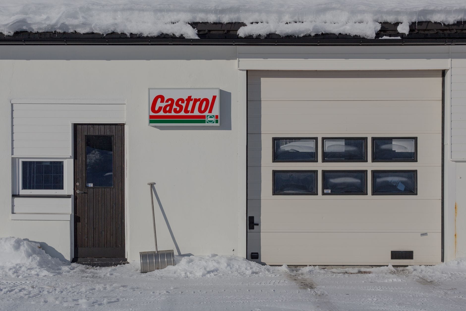 Zaśnieżony podjazd z bramą garażową, łopatą i szyldem Castrol